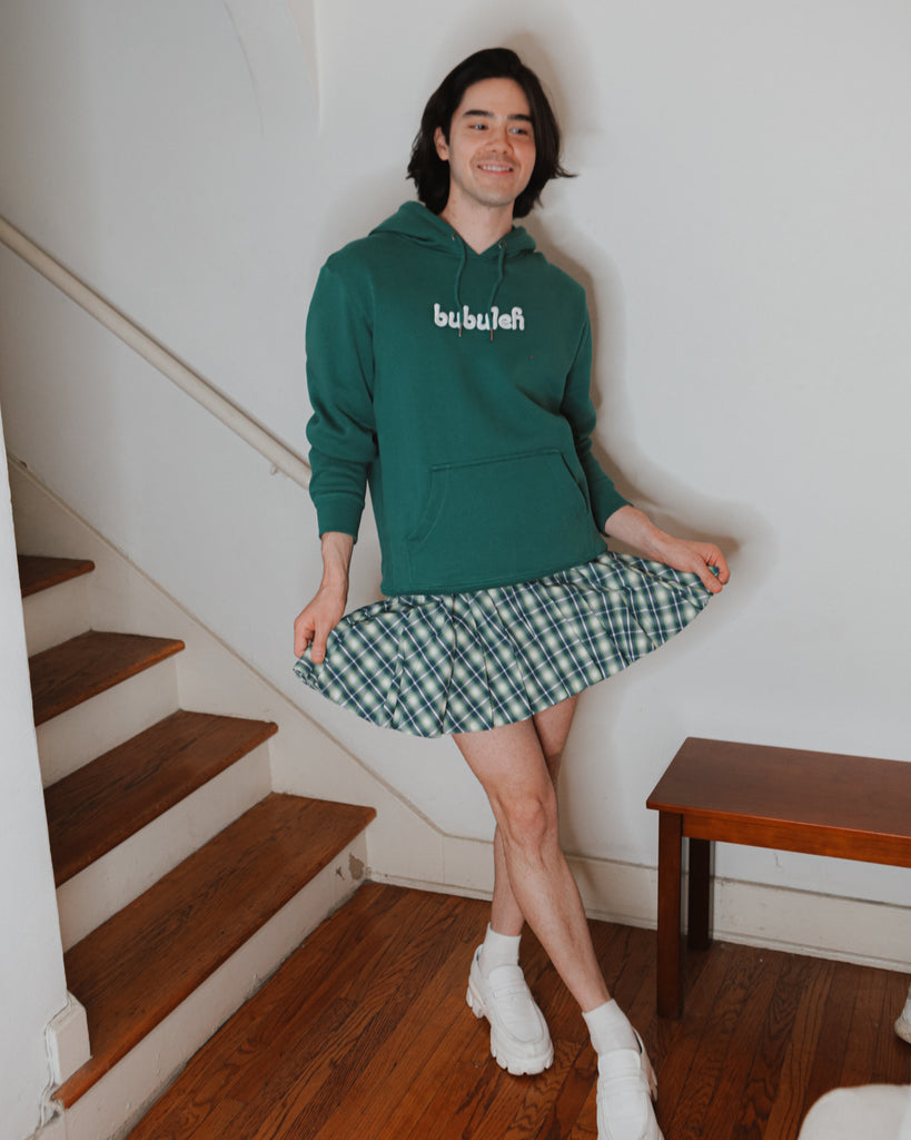 Comedian Jared Goldstein smiling and wearing a green bubuleh hoodie with a matching plaid skirt.